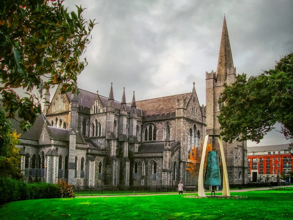 Vista da Catedral St Patrick's, Dublin com  igreja de pedra, que o céu está nublado e cinza e  há um jardim e um belo gramado verde ao redor com um sino próximo a igreja. - Representa chip de celular Dublin