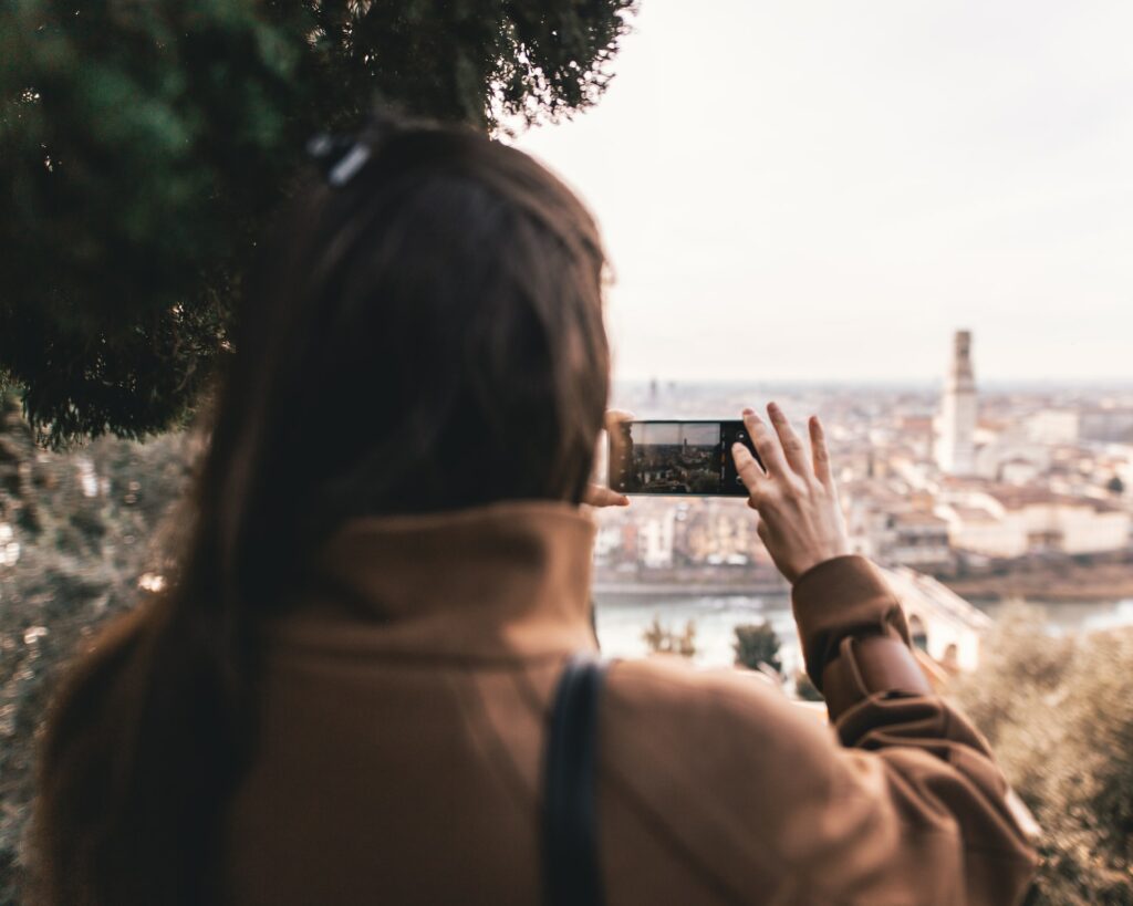 Uma mulher com cabelos longos e usando uma jaqueta marrom está segurando um celular na horizontal tirando uma foto de uma vista de Verona na Itália