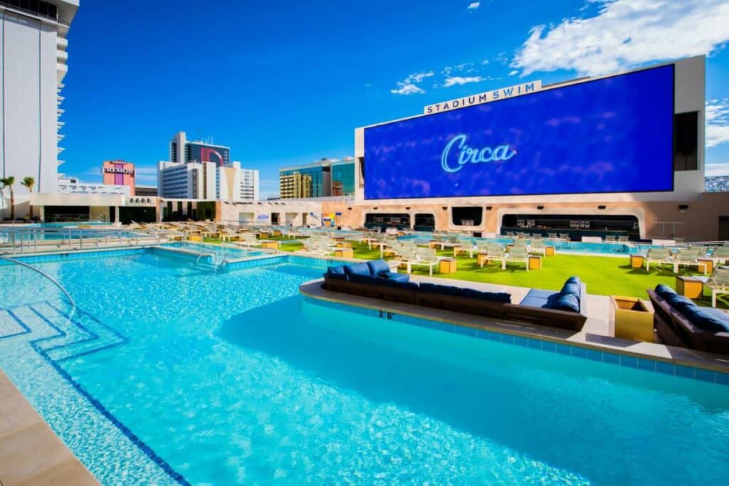 Piscina muito ampla do Circa Resort & Casino - Adults Only, ao lado da piscina, tem um gramado com muitos sofás azuis escuros, almofadas e algumas espreguiçadeiras brancas