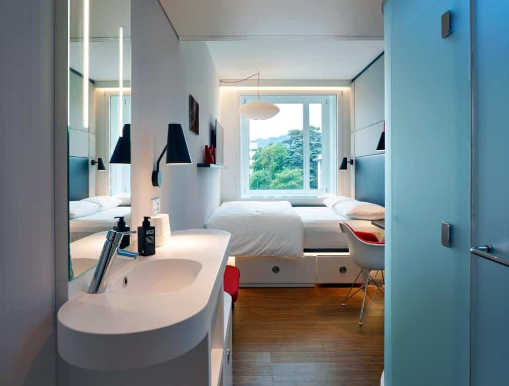 Quarto do citizenM Zürich, um dos hotéis em Zurique, com cama de casal, cadeira, e pia do banheiro com espelho