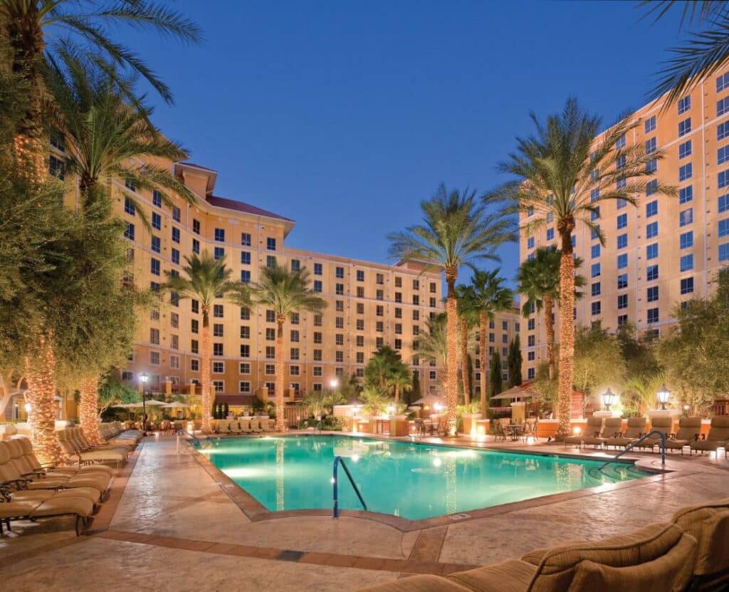 Uma piscina espaçosa no Club Wyndham Grand Desert com os prédios do hotel ao redor, no deck há palmeiras, iluminação e espreguiçadeiras estofadas em bege
