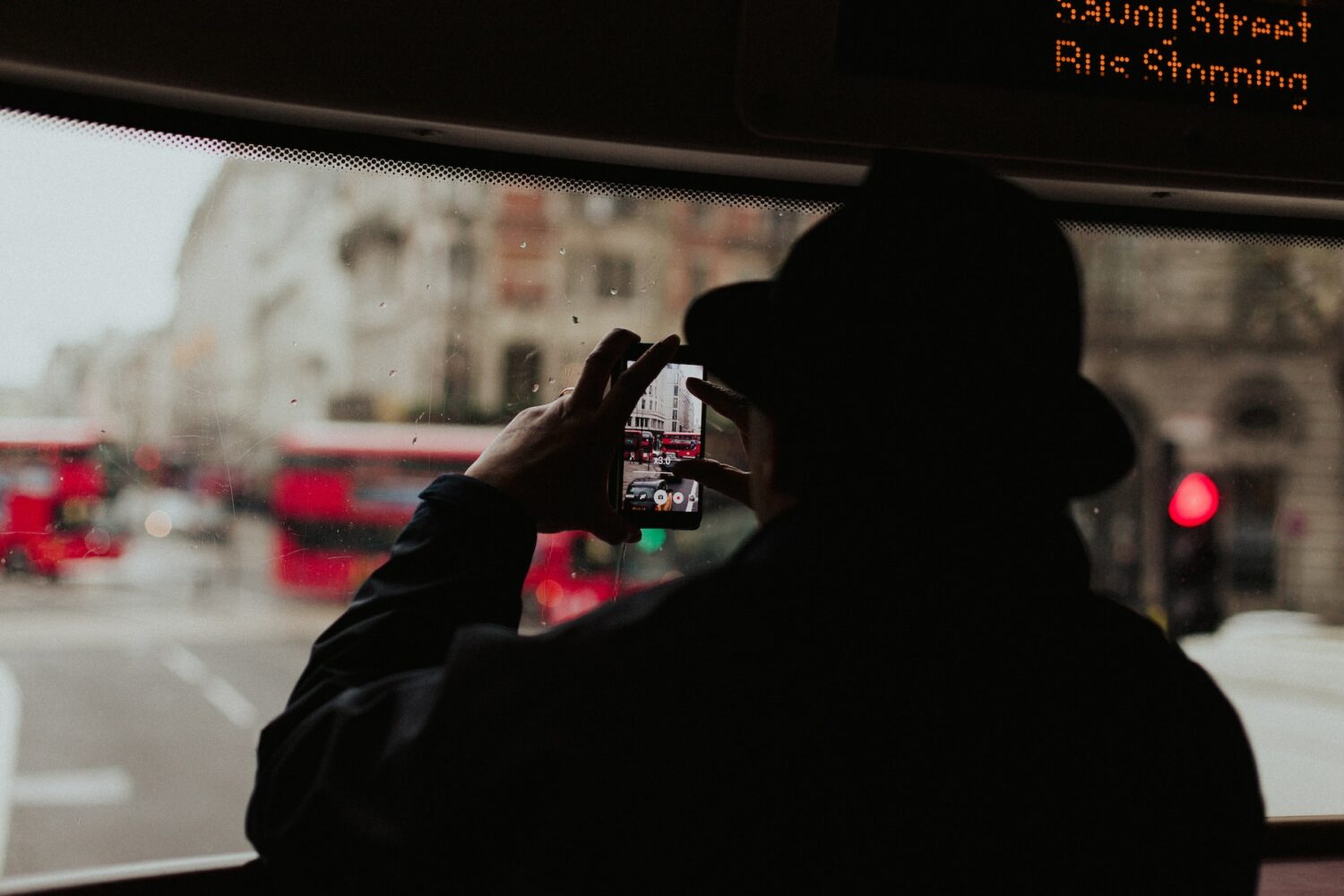Uma mulher de costas usando um casaco e um chapéu, ela está segurando um celular na posição horizontal para fotografar uma rua cheia de ônibus vermelhos