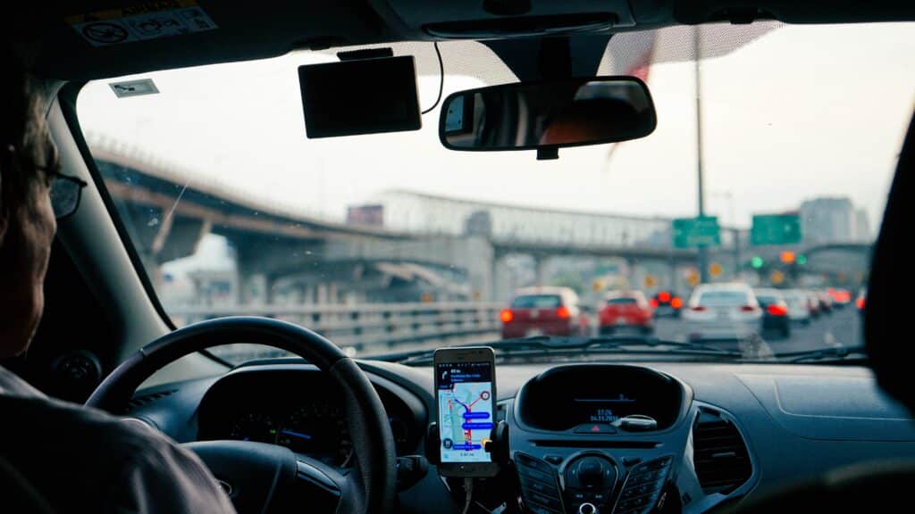 homem de meia idade, usando óculos, dirigindo um carro numa rodovia movimentada, um celular android está preso no painel do carro mostrando as possíveis rotas para seguir