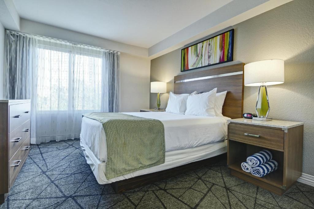Quarto do Desert Rose Resort com uma janela, chão de carpete cinza com desenhos azuis, uma mesa de cabeceira com uma gaveta e toalhas enroladas, um abajur, uma cama de casal do lado direito e, de frente para cama, uma cômoda de madeira com gavetas