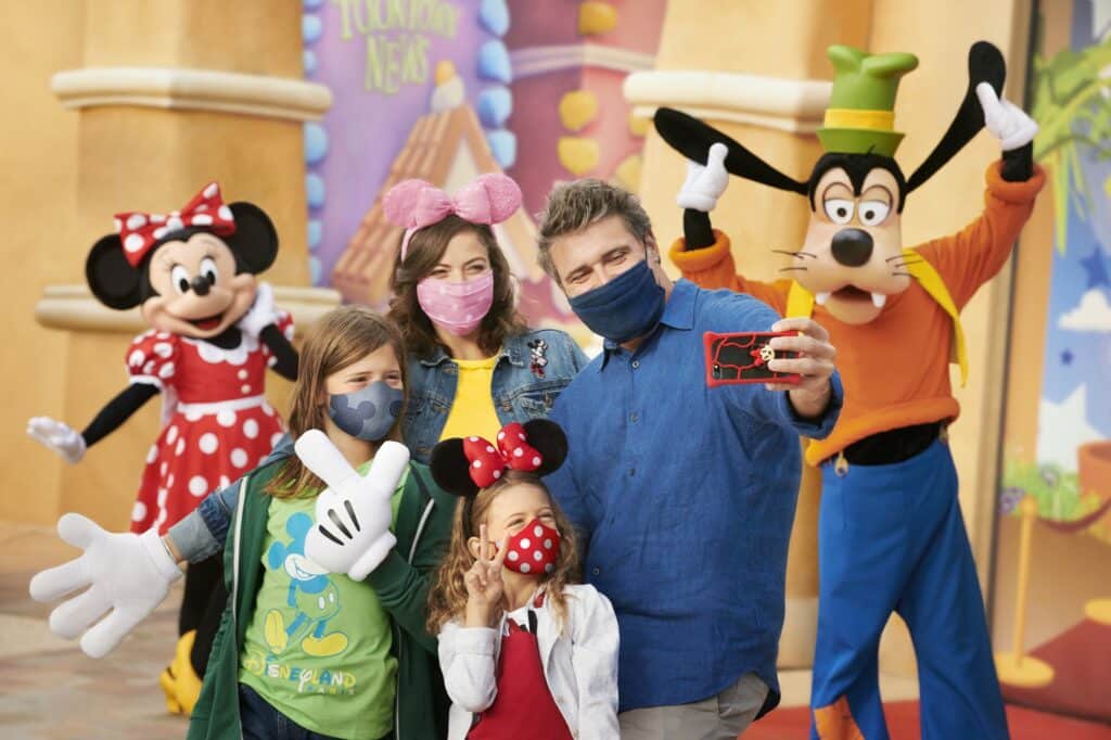 Uma família com um homem, uma mulher, um menino e uma menina, usando alguns acessórios do Mickey e da Minnie, tirando uma selfie com os personagens do Pateta e da Minnie atrás deles, na Disneyland Paris