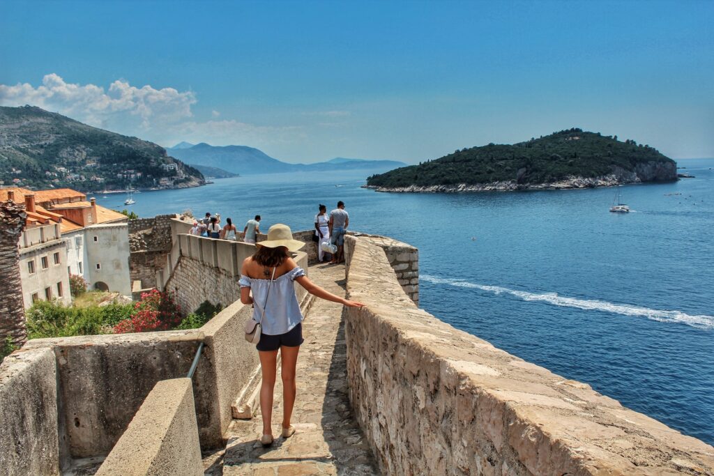 Mulher de chapéu, short curto azul marinho e blusa azul claro, com uma mão em cima de um muro baixo, olhando o mar azulado de Dubrovnik. Há também outros turistas olhando o mar e a paisagem da região