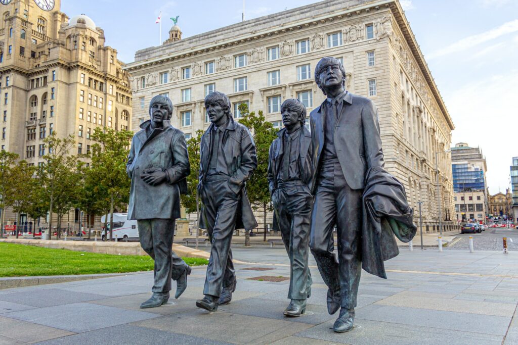 Estátua de bronze dos Beatles, em Liverpool, em uma praça com gramado ao lado, algumas árvores e prédios históricos atrás