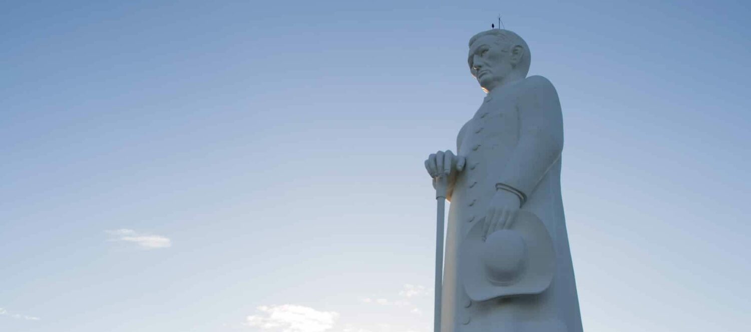 Estátua de Padre Cícero toda branca, um padre de batina com um cajado e o céu azul