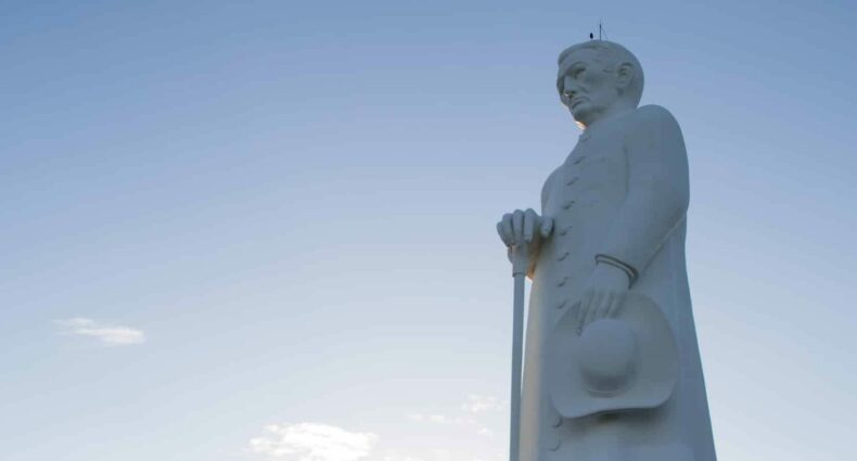 Estátua de Padre Cícero toda branca, um padre de batina com um cajado e o céu azul
