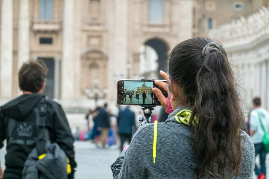 Uma mulher de costas, os cabelos são longos e pretos e estão presos, ela está segurando um celular na horizontal para fotografar um ponto turístico de Roma