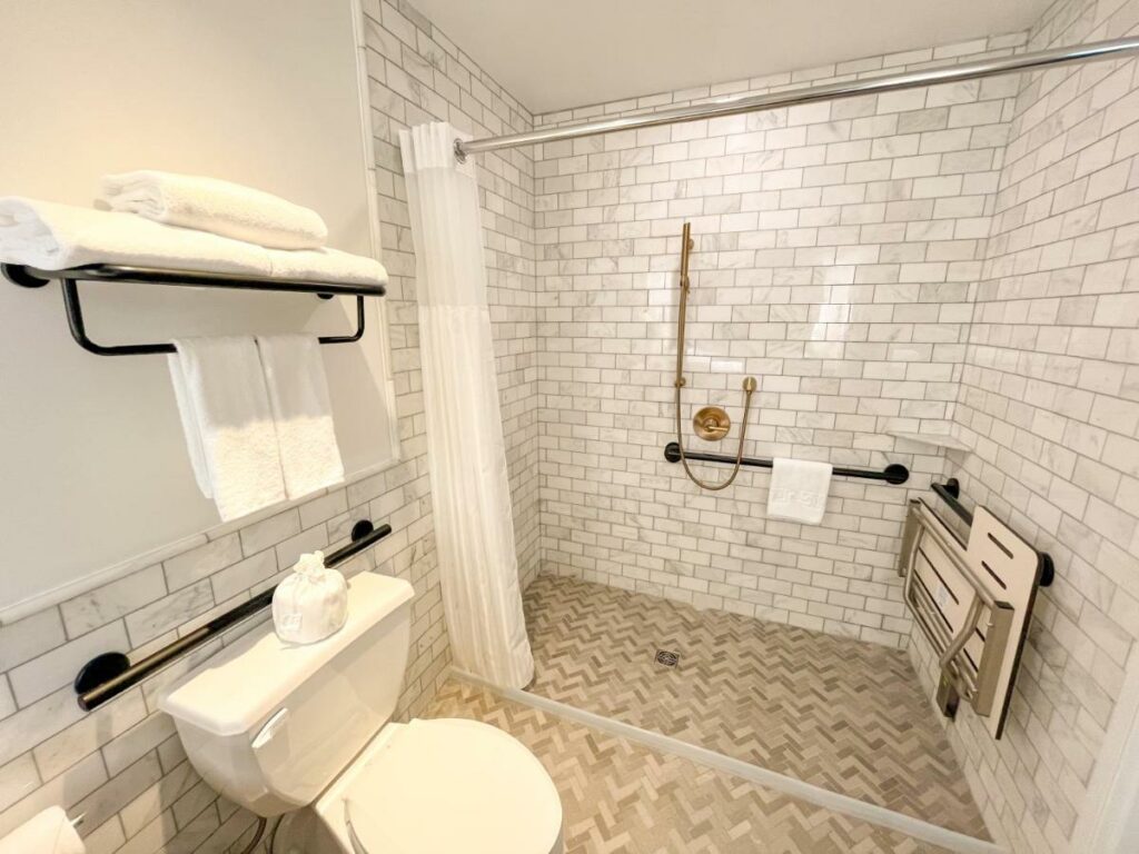 Banheiro no EXchange Hotel Vancouver com um box adaptado com barras de apoio e uma cadeira dobrável, o vaso sanitário do lado esquerdo também conta com barras de apoio