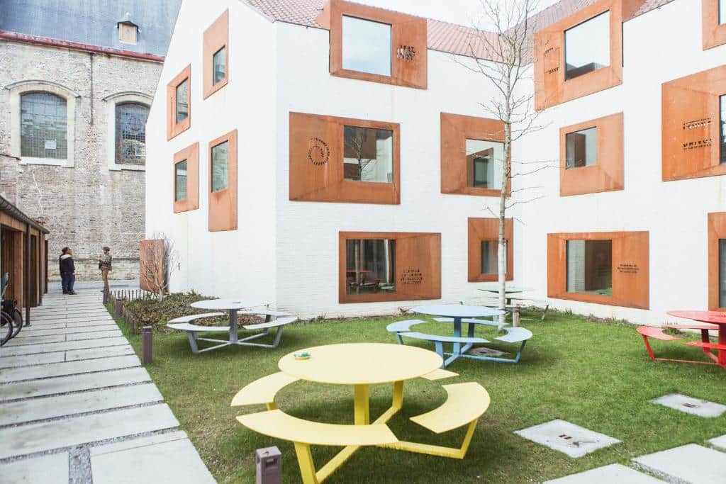 área externa do Snuffel Hostel em Bruges com mesinhas e bancos coloridos com o prédio atrás branco com janelinhas laranjas