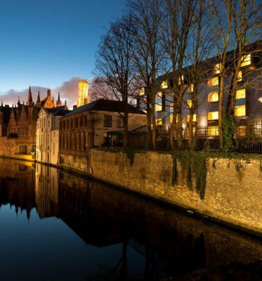 vista da fachada do Grand Hotel Casselbergh Brugge, um dos hotéis em Bruges, na beira do rio, com algumas árvores na frente e outras construções
