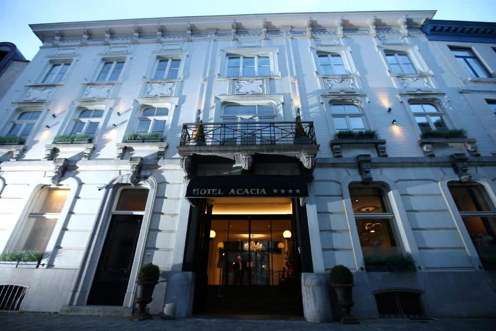 fachada branca em estilo clássico do Hotel Acacia, um dos hotéis em Bruges, com a entrada em portas de vidro e luzes no interior