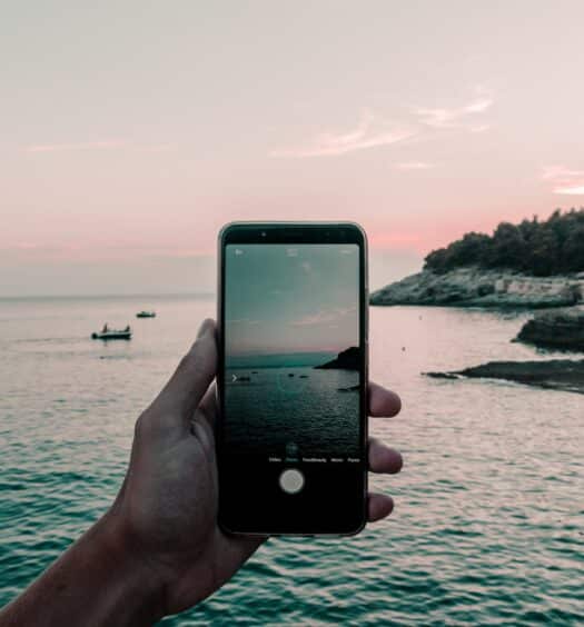 Mão segurando um celular preto, com a tela da câmera aberta mostrando a imagem de uma praia localizada na Croácia