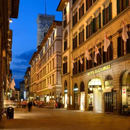 Frente do FH55 Hotel Calzaiuoli, em Florença, sendo o prédio com 4 andares além do térreo, algumas bandeiras enfileiradas no primeiro andar, e várias janelas espalhadas no prédio que fica num calçadão com pessoas andando na frente
