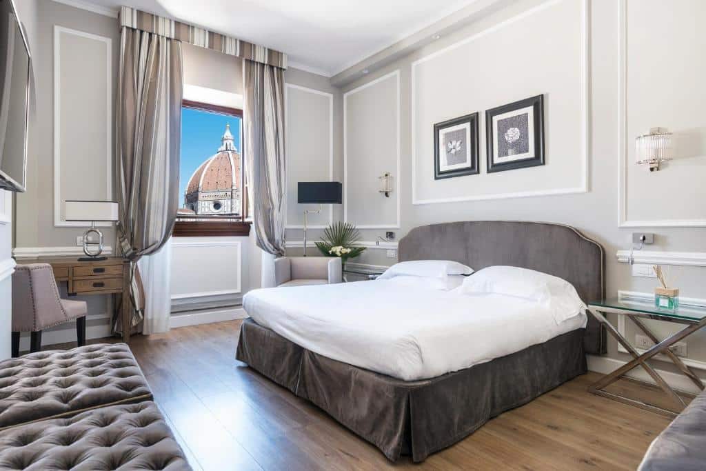 Quarto do FH55 Hotel Calzaiuoli, decorado com cores cinza e branco, contendo uma cama de casal, escraninha nos lados, mesinha com cadeira estofada e janela mostrando a cúpula de uma igreja que fica aem frente ao local