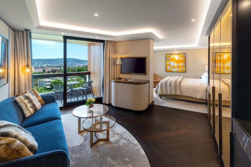 Quarto do FIVE Zurich, de 61 m², com sala de estar com um sofá azul, TV e mesinha redonda ao meio; varanda com mesa, duas cadeiras e vista da cidade com bastante natureza verde; além de parte do quarto com cama macia e quadro com desenho abstrato amarelo