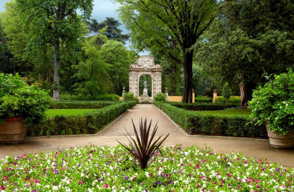 Jardim do Four Seasons Hotel, em Florença, com bastante verde, contando com um pequeno jardim redondo no chão com flores rosas, roxa, verde e branca, um caminho levando em direção a uma estátua, além de gramado verde e árvores altas nos lados
