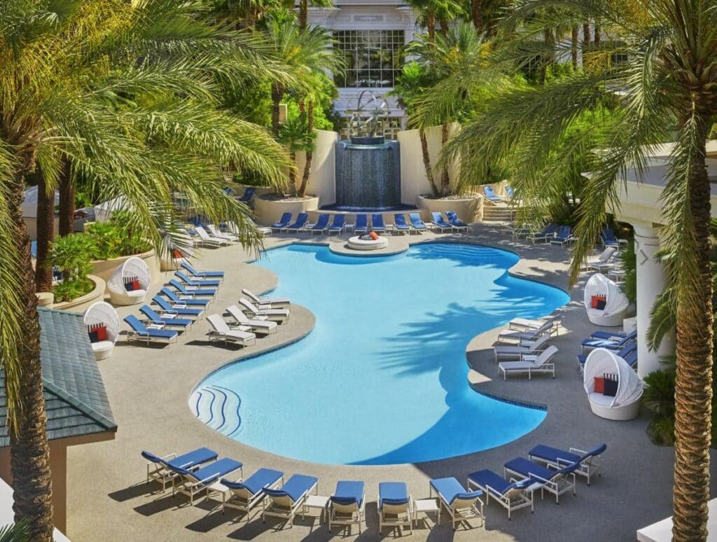 Piscina com bordas arredondadas no Four Seasons Hotel Las Vegas  com um deck ao redor com espreguiçadeiras azuis e algumas poltronas brancas com almofadas, além de muitos coqueiros