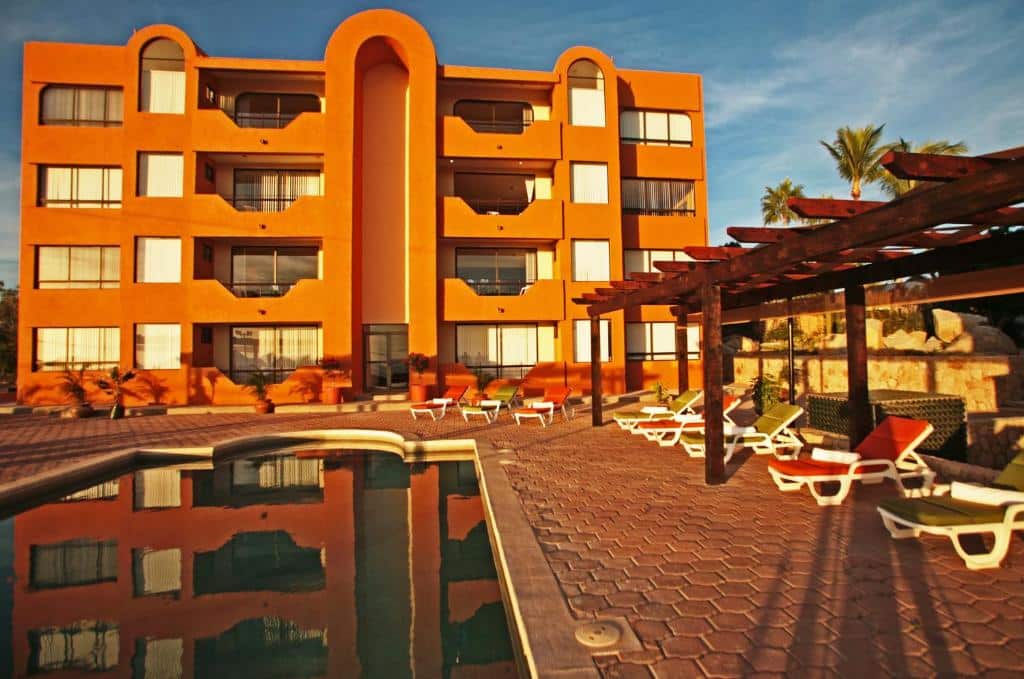 Frente do Sunrock Condo Hotel com cores laranjas e sacadas. Na frente também á uma piscina e cadeiras em volta da piscina. Representa hotéis em Los Cabos.