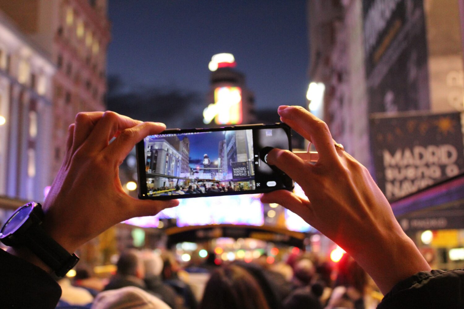 Duas mãos tirando uma foto com o celular, em uma noite iluminada na Gran Via, em Madri, contendo várias pessoas na frente e mostrando a imagem da avenida movimentada na tela