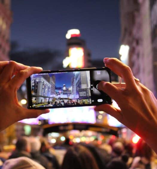 Duas mãos tirando uma foto com o celular, em uma noite iluminada na Gran Via, em Madri, contendo várias pessoas na frente e mostrando a imagem da avenida movimentada na tela