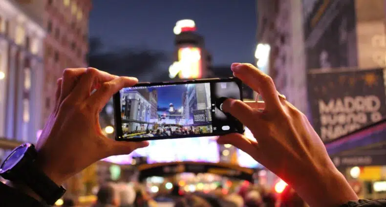 Duas mãos tirando uma foto com o celular, em uma noite iluminada na Gran Via, em Madri, contendo várias pessoas na frente e mostrando a imagem da avenida movimentada na tela