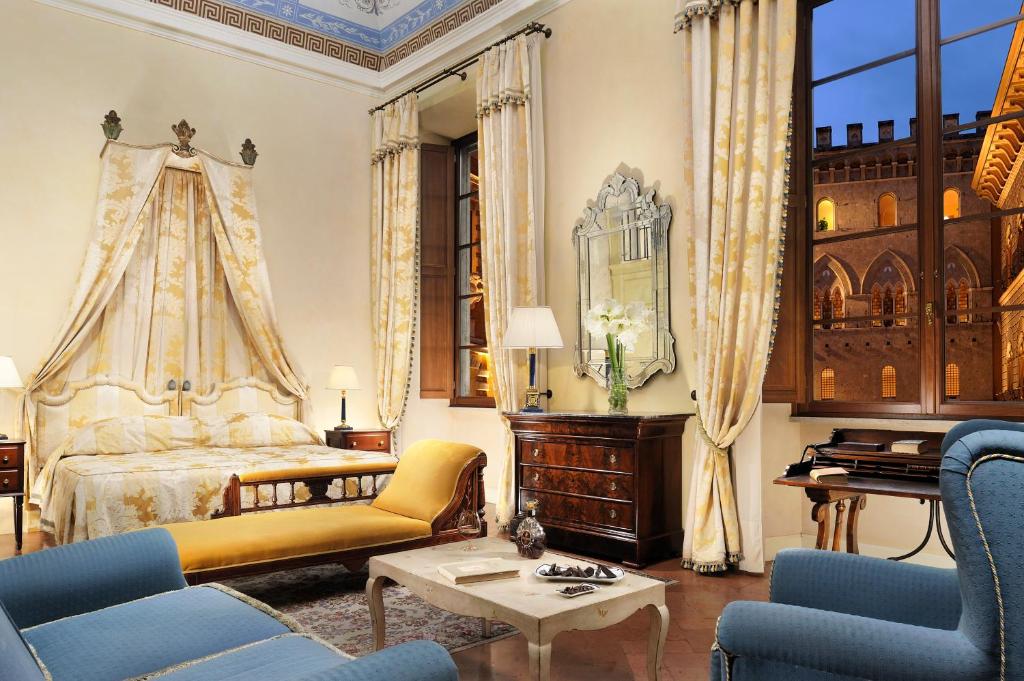 Suíte Heritage do Grand Hotel Continental Siena, de 50 m², com móveis de época, sendo uma cama com colchas combinando com as cortinas, poltronas grandes azuis, espelho, e janelas mostrando um edifício parecido com um castelo