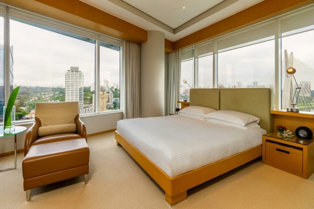 Quarto no Grand Hyatt São Paulo com uma cama de casal, duas janelas com cortinas, duas mesinhas de cabeceira de madeira com abajures, além de uma poltrona de couro marrom, para representar hotéis perto do Autódromo de Interlagos