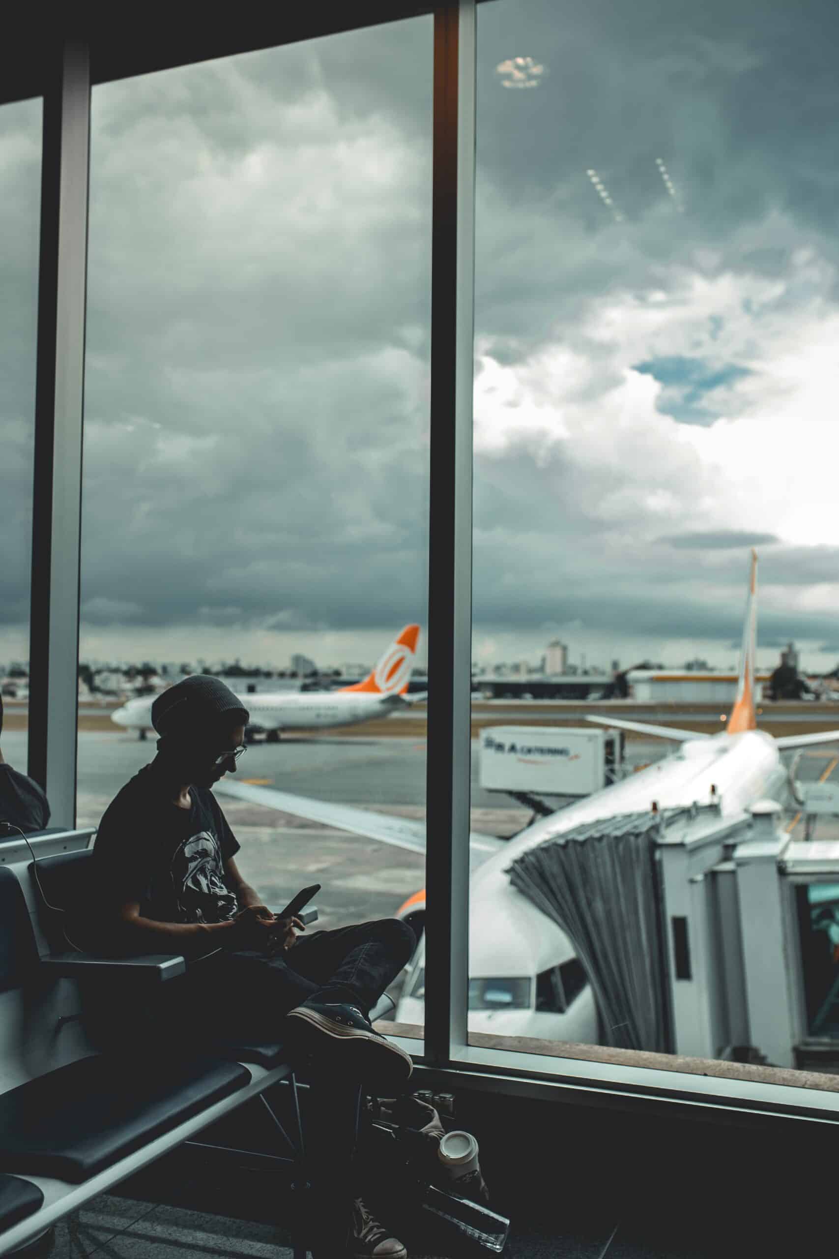 jovem homem sentado à esquerda da imagem no aeroporto de interlagos, bem em frente as largas janelas do aeroporto com vista para alguns aviões da companhia Gol