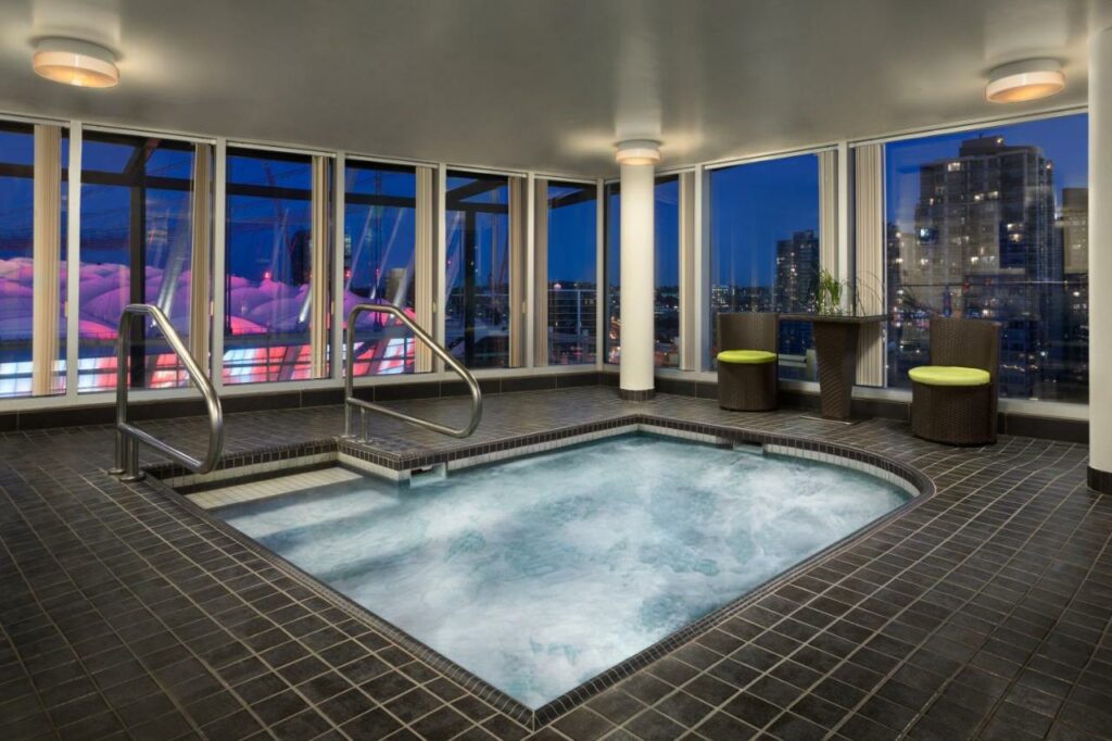 Piscina coberta do Hampton Inn & Suites, by Hilton - Vancouver Downtown com janelas amplas ao redor dando vista para a cidade, a piscina é pequena e o deck ao redor conta com lugares para se sentar