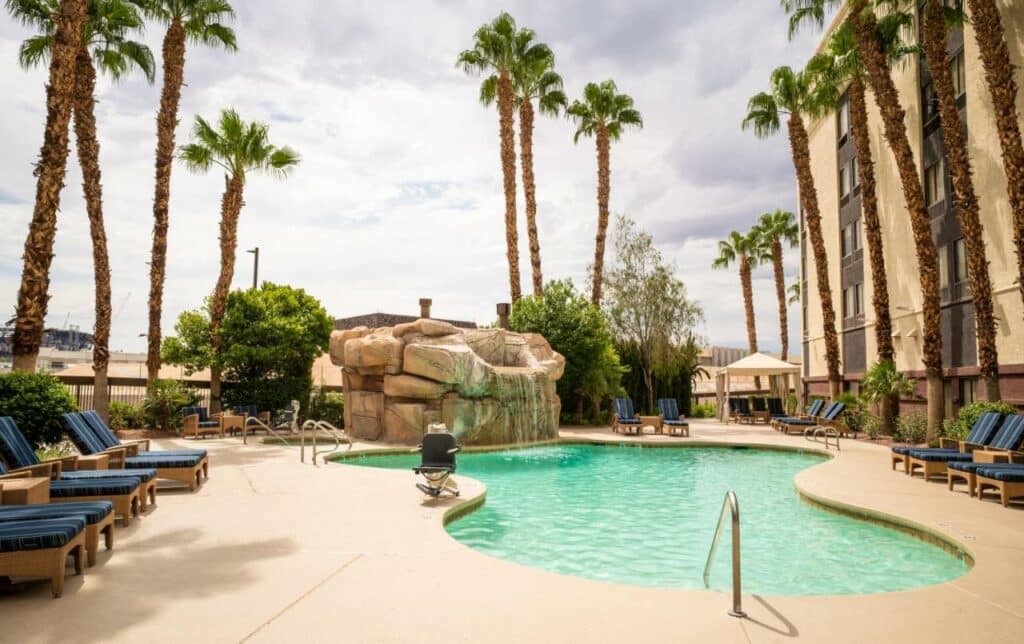 Piscina do Hampton Inn Tropicana Las Vegas com uma ampla rocha que faz uma pequena cascata para dentro da piscina, ao redor da piscina há um deck com espreguiçadeiras azuis, e muitos coqueiros