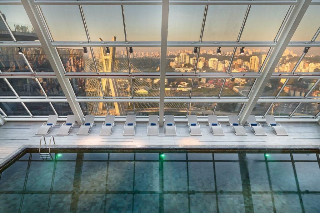 Piscina coberta do Hilton Sao Paulo Morumbicom vista direta para a ponte estaiada de São Paulo, no deck há algumas espreguiçadeiras com as janelas de vidro dando vista para a cidade
