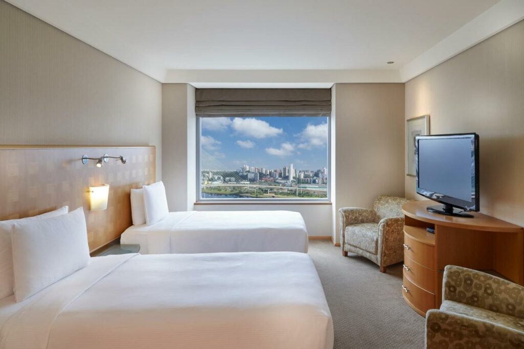 Quarto no Hilton Sao Paulo Morumbi com uma janela ampla com vista para a cidade, há duas camas de solteiro, duas poltronas, e um móvel de madeira com uma televisão