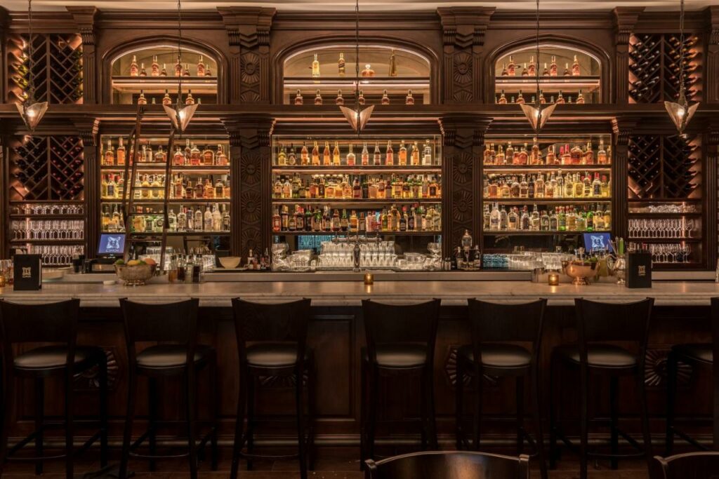 Bar interno do Hollywood Historic Hotel em estilo de época, com tudo montado em tons de madeira escura, um balcão com seis lugares, na parte do bar as garrafas das bebidas ficam expostas em uma espécie de vitrine