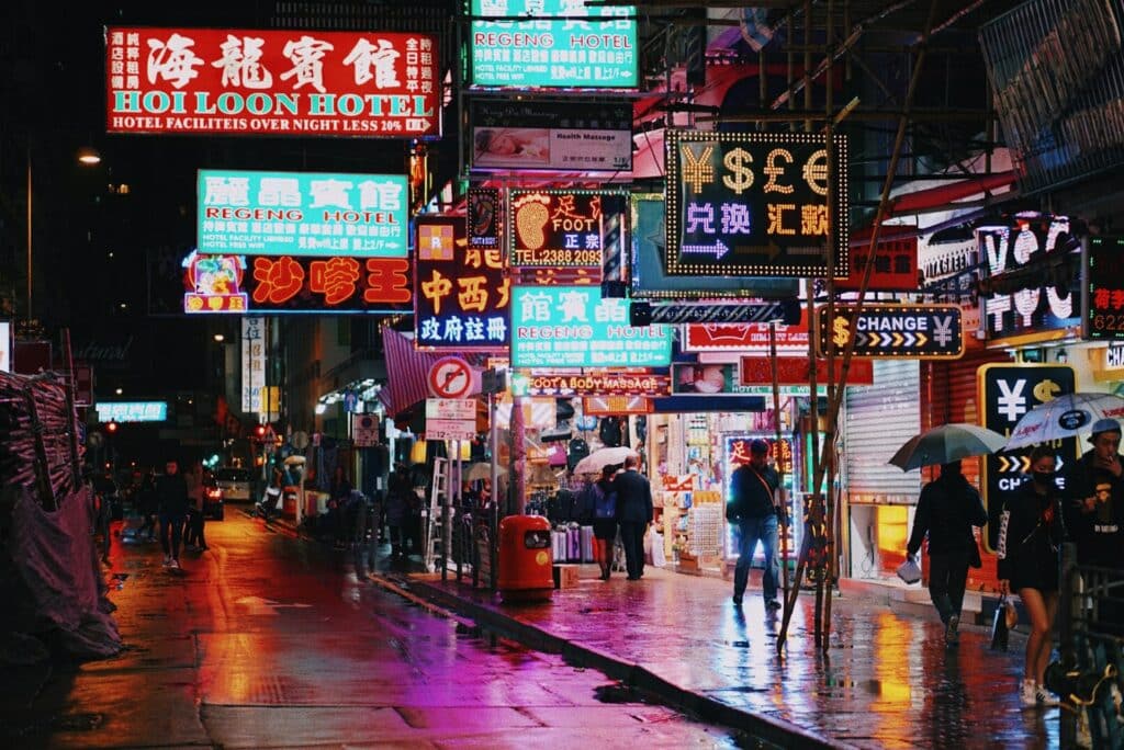 Rua de Hong-Kong em noite chuvosa cheia de comércios e hotéis iluminados com neon e luzes coloridas, pessoas circulam com guarda-chuva para representar o seguro viagem Hong Kong.