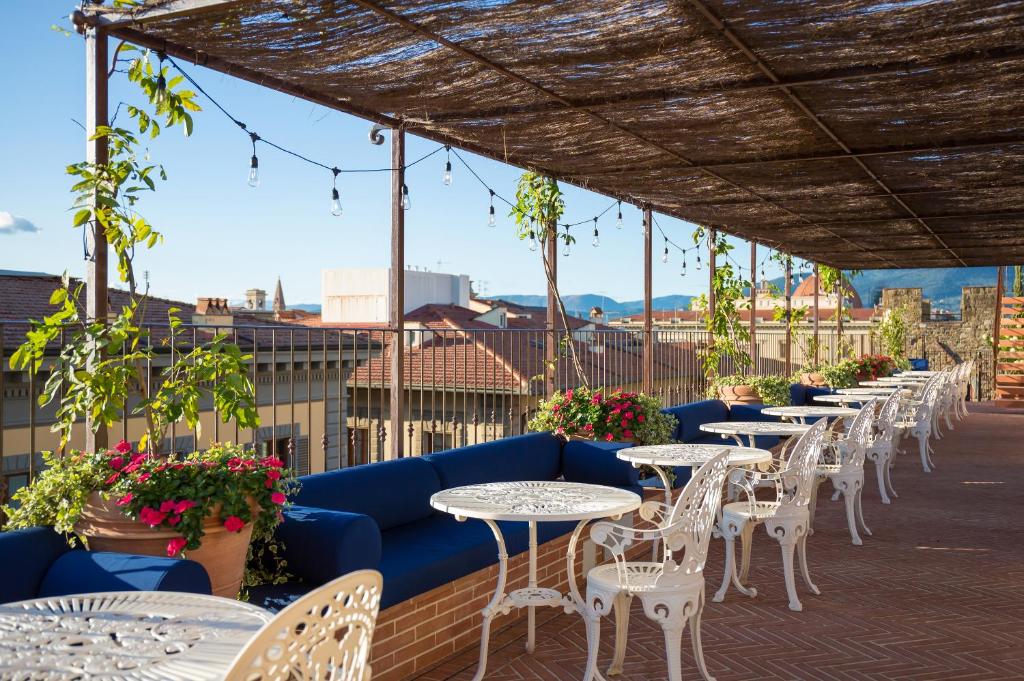 Varanda do Hotel Calimala, em Florença, com várias mesas redondas brancas enfileiradas, com uma cadeira branca combinando com a mesa de um lado, poltronas azuis de outro, flores verdes e rosa enfeitando o espaço, e vista para algumas construções da cidade