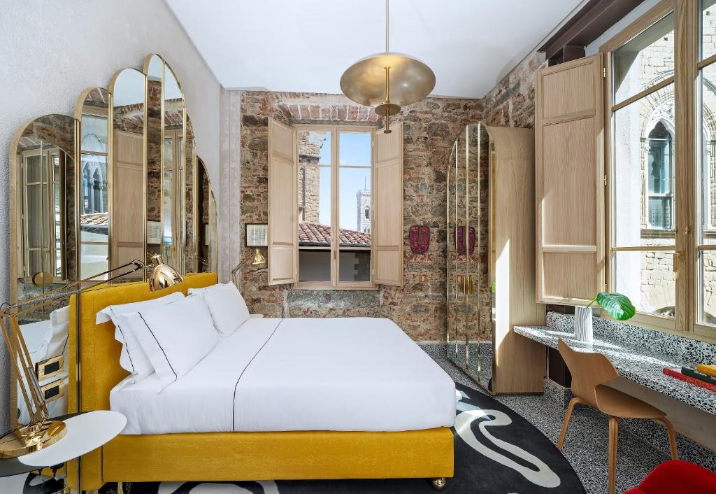 Quarto duplo superior do Hotel Calimala, de 15 m², com uma cama de casal branca e amarela macia, cabeceira espelhada encostada na parede, janelas grandes de madeira mostrando prédios vizinhos, um guarda-roupa espelhado combinando com a cabeceira e um balcão com uma cadeira
