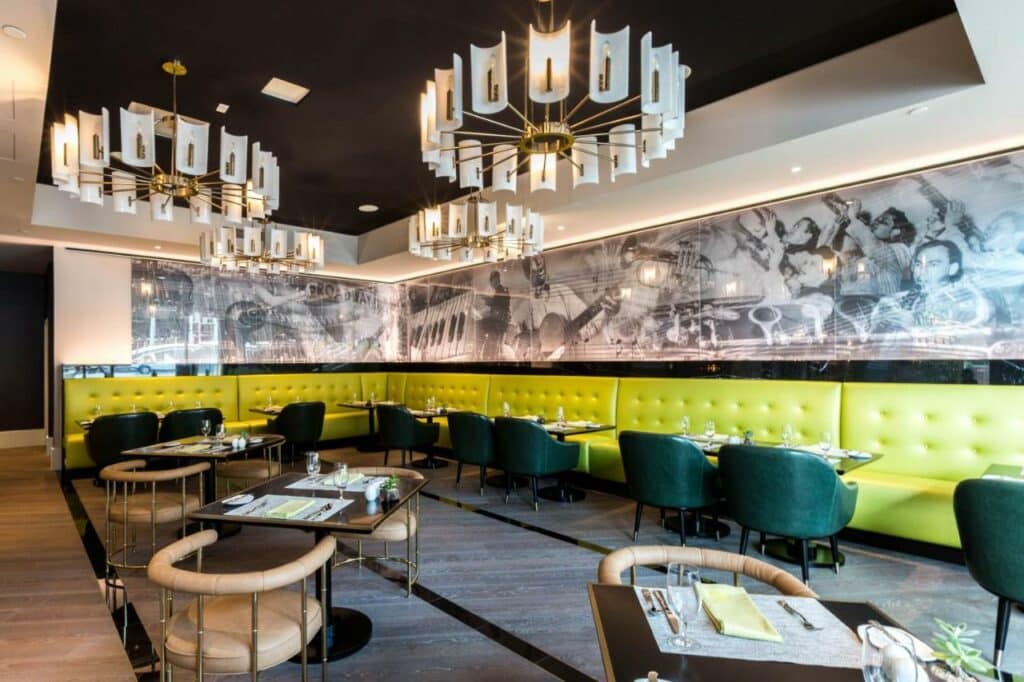 Restaurante do Hotel Indigo - Los Angeles Downtown, an IHG Hotel com muitos sofás, mesas e poltronas para se sentar, as poltronas são verdes escuras e os sofás verde claro, há um painel com fotografias em preto e branco ao redor do ambiente e no texto várioas lustres
