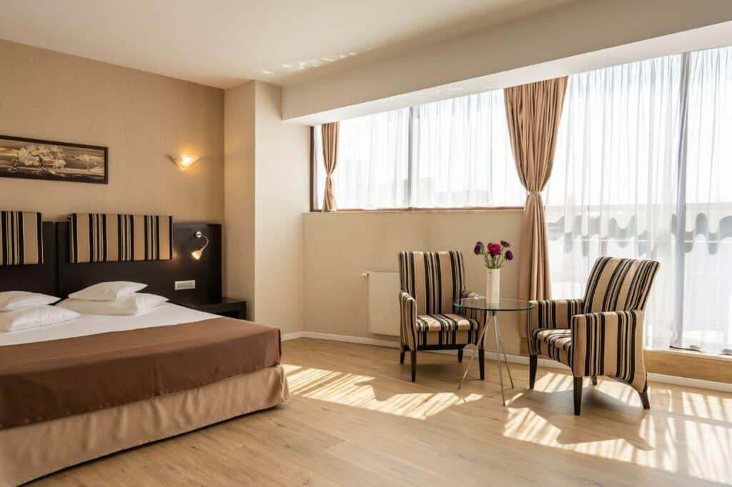 Quarto de hotel, com cama com travesseiros e cobertor, mesa de vidro com cadeiras em volta, um vaso de flor ao centro, janela e sacada com cortinas transparentes.