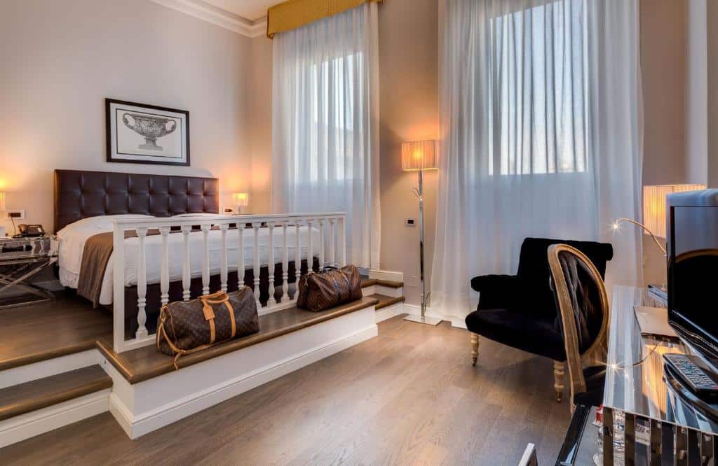 Quarto do Hotel Roma, de 22 m², com uma cama de casal, poltrona preta, mesa com uma cadeira e um monitor em cima, chão de madeira e duas janelas. Há degraus entre a cama e mesa