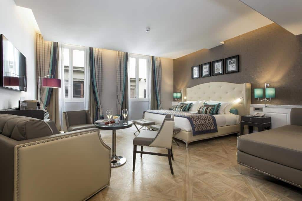 Quarto espaçoso do Hotel Spadai, um dos hotéis em Florença, com cama de casal grande, um abajur verde de cada lado da cama, sofá ao lado cinza, e uma pequena área de estar integrada com mesa redonda, duas cadeiras, uma poltrona grande e TV