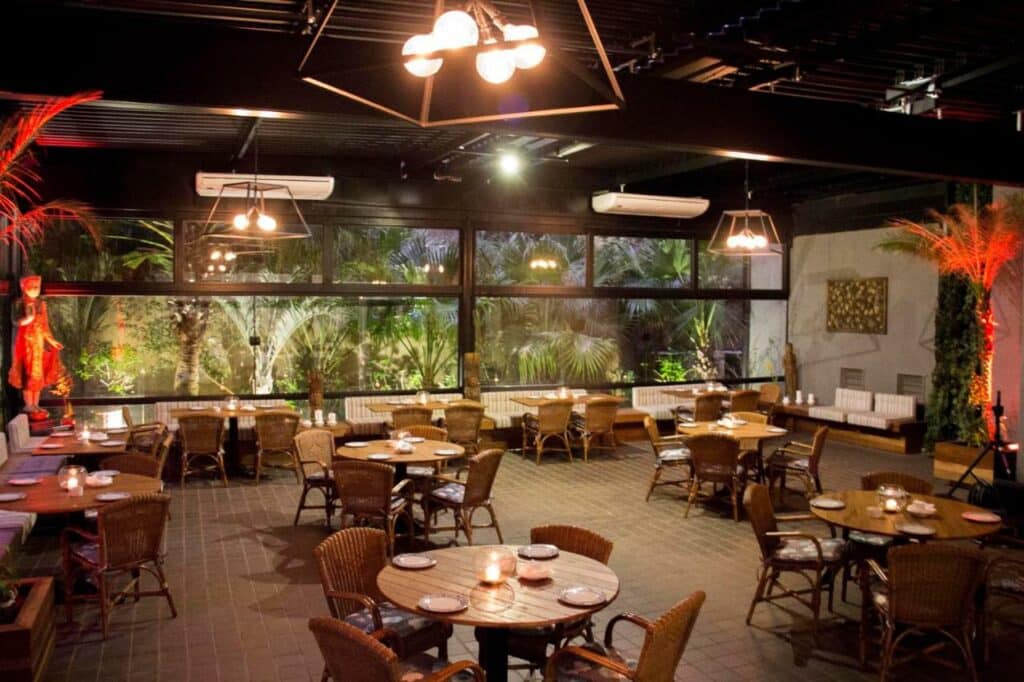 Área para refeições no Hotel Transamerica Berrini com janelas amplas e de vidro que dão vista para o jardim, o chão é de madeira, assim como as mesas redondas e as cadeiras, para representar hotéis perto do Lollapalooza
