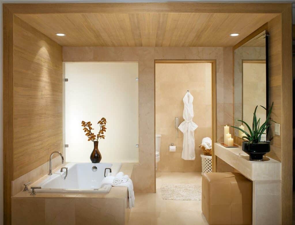 Banheiro muito amplo do Huntley Santa Monica Beach com uma banheira em um dos ambientes com um espelho e algumas levas, depois há uma porta que leva para o sanitário e a pia, com um roupão pendurado, todo o ambiente é em mármore e madeira clara