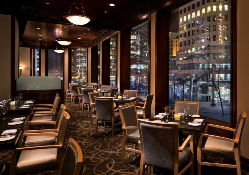 Área do restaurante do Hyatt Regency Vancouver  com muitas mesas e cadeiras de madeira, o local tem amplas janelas viradas diretamente para a rua, o carpete do lugar é cinza com desenhos circulares brancos, no teto há luminárias brancas redondas