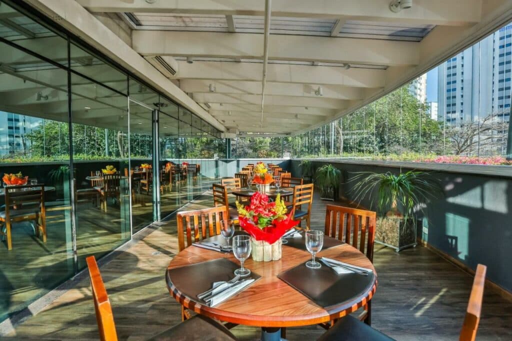 Área para refeições com vista para a cidade no ibis Sao Paulo Congonhas com mesas de madeira redondas e quatro cadeiras de madeira, há arranjos de flores, taças e talheres sob as mesas, para representar hotéis perto do Autódromo de Interlagos
