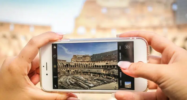 Uma moça segurando um celular na horizontal enquanto fotografa um dos monumentos históricos de Roma com um celular branco