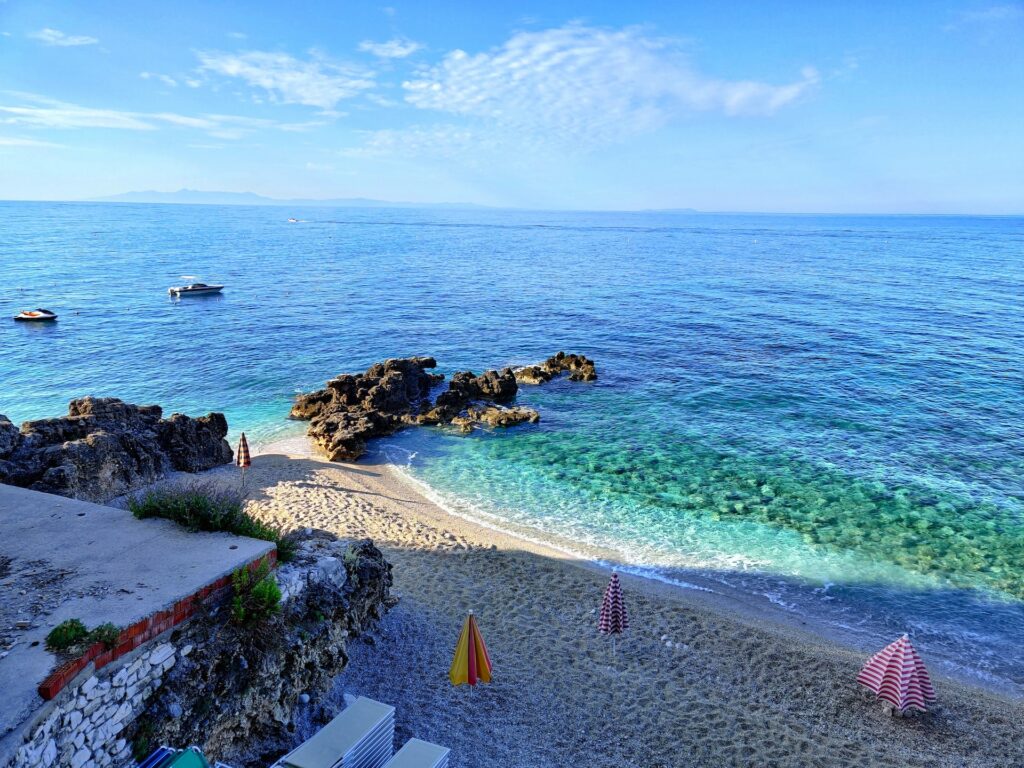 Uma praia em Dhërmi na Albânia, o mar está em tons de verde e azul claro, a faixa de areia é pequena e tem alguns guarda-sóis presos, ao redor da praia, há muitas rochas, assim como dentro do mar próximo da areia