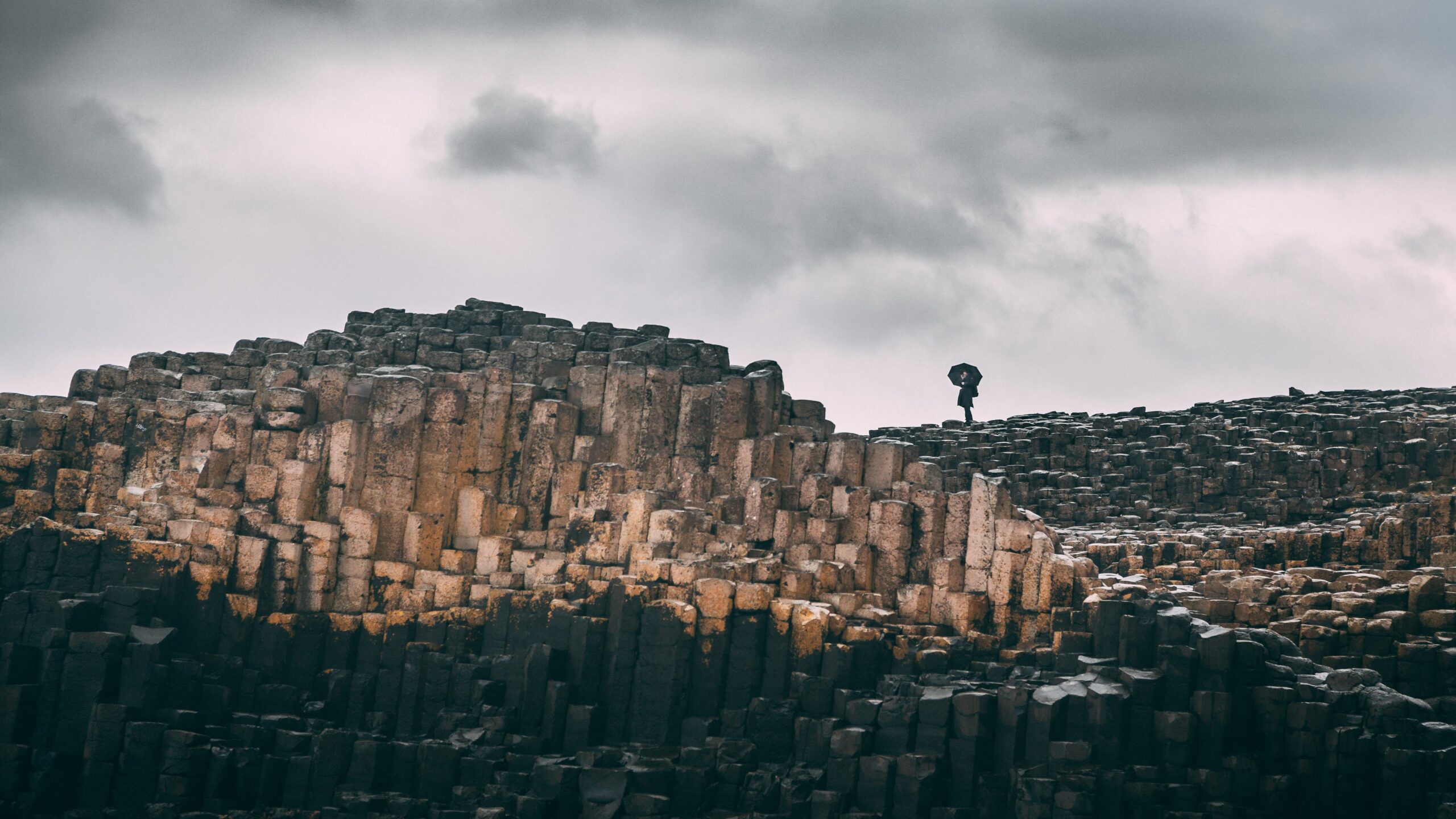céu nublado sobre a estrutura hexagonal de pedra nomeada calçada dos gigantes que fica na irlanda do norte. Ao fundo é possível ver uma mulher parada com um guarda-chuva preto aberto.